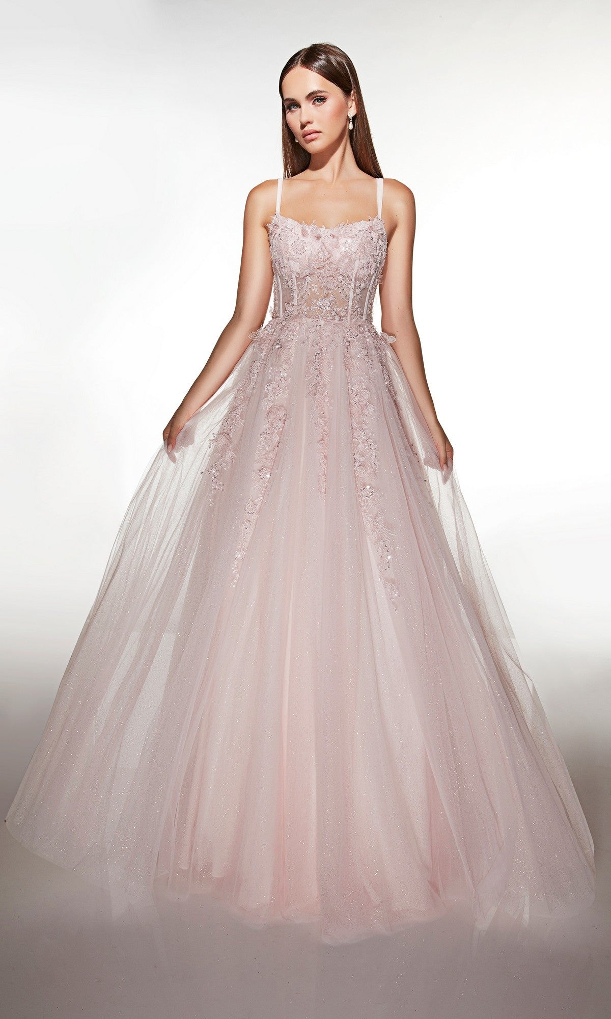 A woman models the Alyce 62203 long prom dress, featuring light pink tulle, a fitted corset bodice with thin straps, 3D floral appliques, and a full floor-length skirt against a plain background.